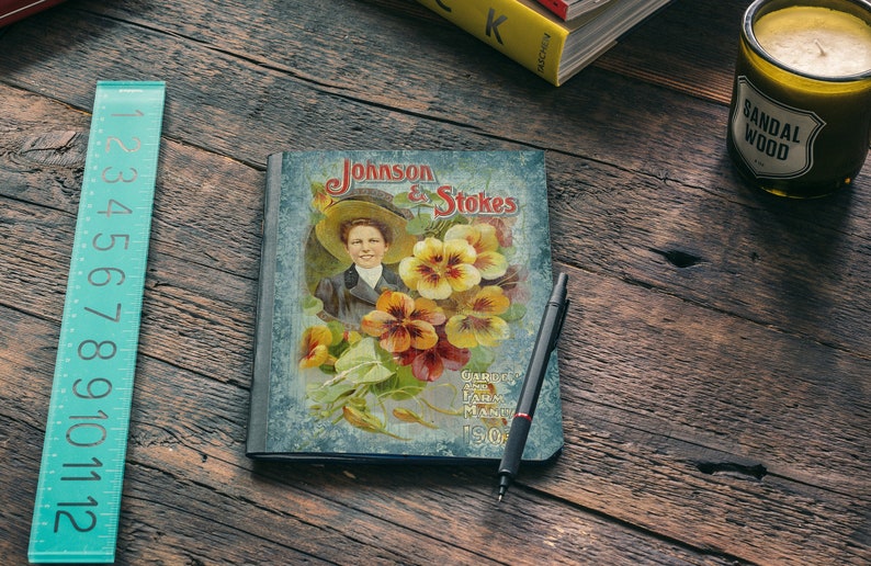 Puede incluir: Un cuaderno de estilo vintage con una cubierta azul que presenta a una mujer con un sombrero y un dise&ntilde;o floral. La cubierta dice "Johnson & Stokes" y "Garden, Farm and Flower Seeds 50c".