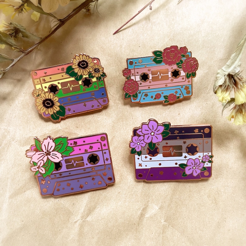 May include: Four enamel pins featuring a cassette tape design with floral accents. Each pin has a different color scheme representing the colors of the LGBTQ+ pride flag. The pins are on a beige background.