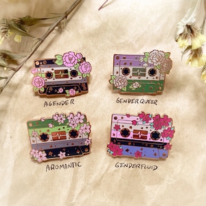 May include: Four enamel pins shaped like cassette tapes with floral accents and text below each pin. The pins represent different identities: Agender, Genderqueer, Aromantic, and Genderfluid.