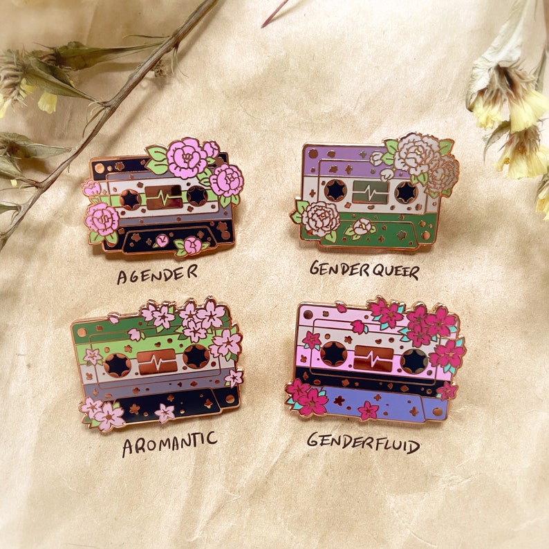 May include: Four enamel pins shaped like cassette tapes with floral accents and text below each pin. The pins represent different identities: Agender, Genderqueer, Aromantic, and Genderfluid.