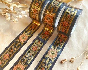 May include: Decorative washi tape rolls in black with gold and red floral and geometric patterns. The tape features sunflowers, starbursts, and diamond shapes. The tape is unrolled, showing the repeating design. The rolls are on a neutral background.
