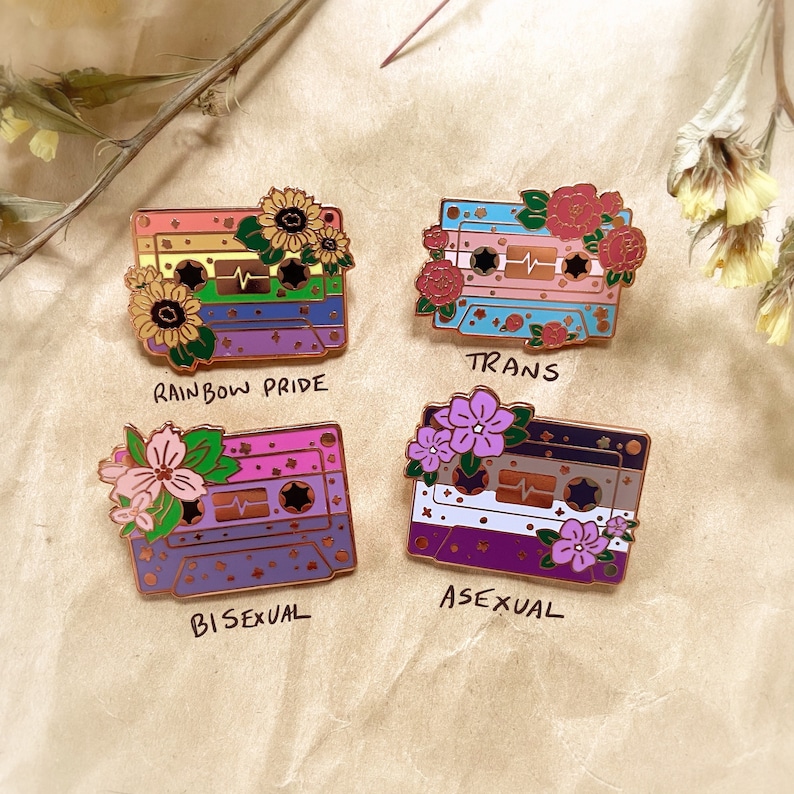 May include: Four enamel pins shaped like cassette tapes with different colored stripes and flowers. The pins represent different sexual orientations: rainbow pride, trans, bisexual, and asexual.
