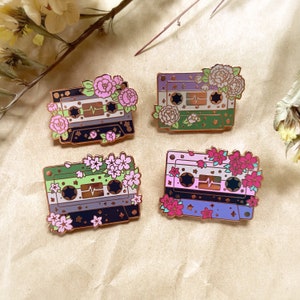 May include: Four enamel pins featuring a cassette tape design with floral accents. Each pin has a different color scheme: green and white, purple and white, pink and white, and blue and white.