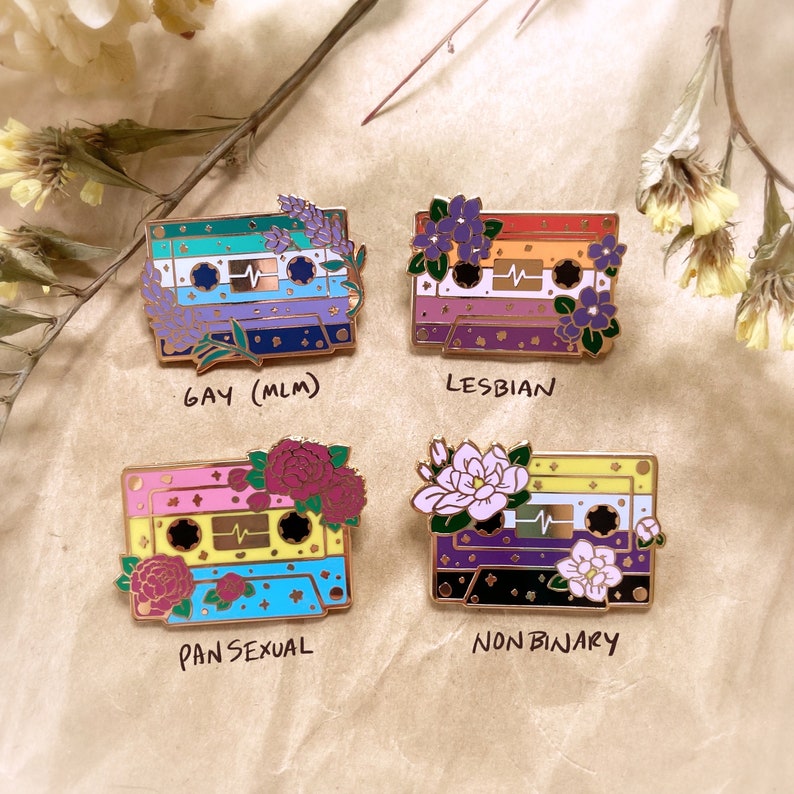May include: Four enamel pins shaped like cassette tapes with floral designs and text below each pin. The pins represent different sexual orientations: Gay (MLM), Lesbian, Pansexual, and Non-Binary.