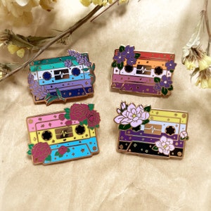 May include: Four enamel pins shaped like cassette tapes with floral accents and a heartbeat symbol. Each tape features a different pride flag: green, blue, and white; red, orange, yellow, and purple; pink, yellow, and blue; and black, purple, white, and yellow.