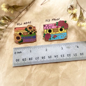 May include: Two cassette tape pins with floral accents. The pin on the left is labeled "NEW MANU" and features a rainbow color scheme with sunflowers. The pin on the right is labeled "OLD MANU" and features a blue, yellow, and pink color scheme with red roses. Both pins are approximately 1 inch in size.