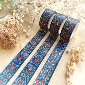 May include: Three rolls of decorative washi tape with a dark blue background and a floral design in red, gold, and light blue. The tape is unrolled, revealing the pattern. The background is a light brown.