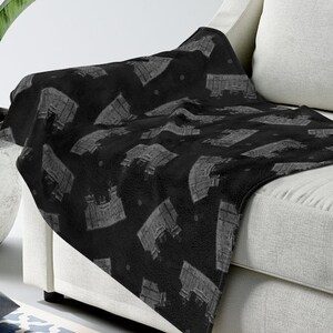May include: A black fleece throw blanket with a white repeating pattern of a building. The blanket is draped over a white couch.
