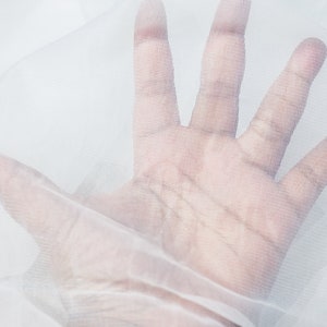 以下が含まれることがあります： 細かいメッシュのテクスチャを持つ白い透き通った生地。生地の上に手が置かれ、メッシュパターンが表示されています。