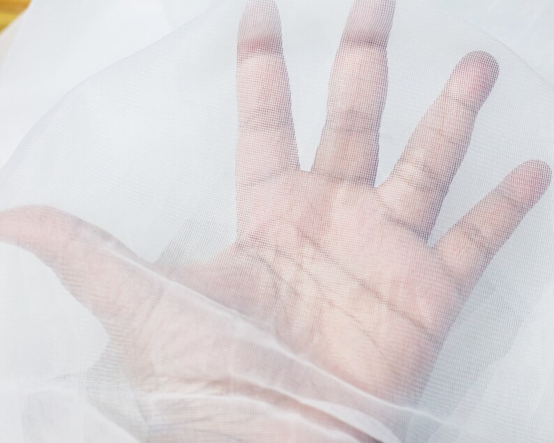 以下が含まれることがあります： 細かいメッシュのテクスチャを持つ白い透き通った生地。生地の上に手が置かれ、メッシュパターンが表示されています。