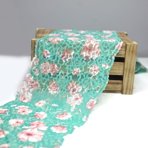 May include: Teal lace trim with a floral pattern in pink and white. The lace is displayed on a wooden crate.