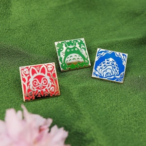 May include: Three enamel pins featuring characters from the animated film 'My Neighbour Totoro'. The pins are square shaped and have a white outline. The first pin features a green Totoro with a white belly. The second pin features a red Totoro with a white belly. The third pin features a blue Totoro with a white belly.