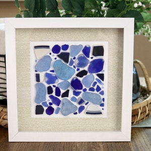 May include: A framed mosaic artwork featuring various shades of blue and white sea glass pieces arranged in a random pattern on a white background.