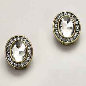 以下が含まれることがあります： 2つの楕円形のクリップイヤリング。各イヤリングは、中央に大きな透明なファセットストーンがあり、小さな透明なストーンのリングで囲まれています。セッティングは金色の金属です。イヤリングは白い背景に表示されています。