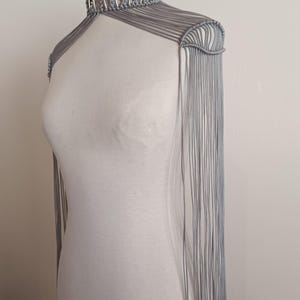 Könnte beinhalten: Ein graues Makramee-Schulterstück mit hohem Kragen und Fransen. Der Kragen hat silberfarbene Ringe. Das Schulterstück wird auf einer weißen Schaufensterpuppe vor einem schlichten weißen Hintergrund präsentiert. Die Fransen hängen vorne und hinten herunter.