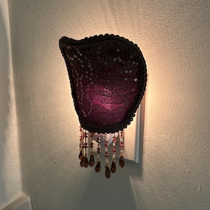 Puede incluir: Una luz de noche decorativa de pared con una pantalla de lentejuelas de color púrpura oscuro. La pantalla está adornada con un material oscuro similar a una cuerda y tiene detalles de cuentas colgantes en tonos marrones y rojos. La luz está enchufada a una toma de corriente blanca.