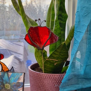 May include: A red stained glass butterfly with black accents sits in a pot of green snake plant. The pot is brown with a textured surface.