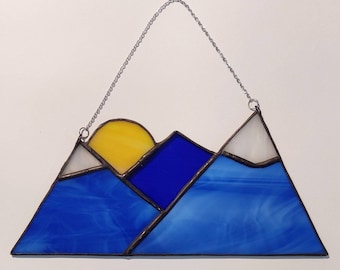 May include: A stained glass sun setting behind a mountain range. The sun is yellow, the mountains are blue and white.