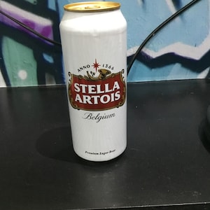 May include: A white aluminium can of Stella Artois beer. The can features a red and gold label with the Stella Artois logo and the words "Anno 1366" and "Belgium". The can also states "Premium Lager Beer".