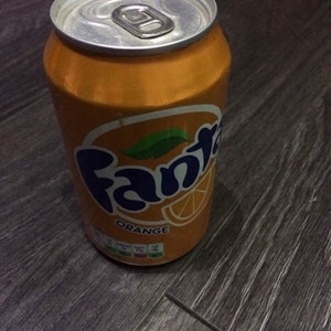 May include: An orange Fanta can with a silver lid. The can displays the Fanta logo in blue and white, with an orange slice graphic. The word "ORANGE" is below the logo. The can is on a dark wood-effect surface.