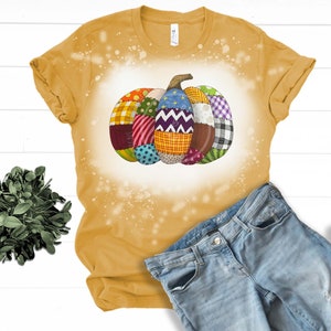 K&ouml;nnte beinhalten: Ein gelbes T-Shirt mit einem Design aus Bleichmittelspritzern, das einen Patchwork-K&uuml;rbis zeigt. Der K&uuml;rbis besteht aus verschiedenen Stoffen in verschiedenen Farben, darunter Rot, Blau, Gr&uuml;n und Gelb.