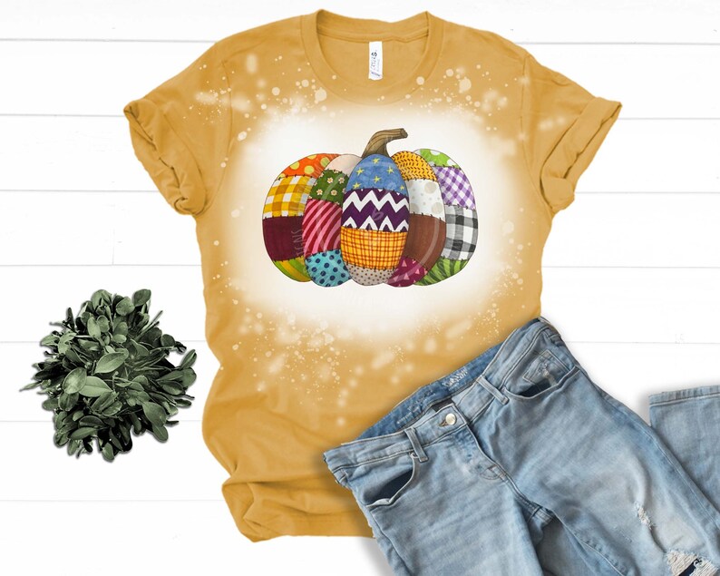 K&ouml;nnte beinhalten: Ein gelbes T-Shirt mit einem Design aus Bleichmittelspritzern, das einen Patchwork-K&uuml;rbis zeigt. Der K&uuml;rbis besteht aus verschiedenen Stoffen in verschiedenen Farben, darunter Rot, Blau, Gr&uuml;n und Gelb.