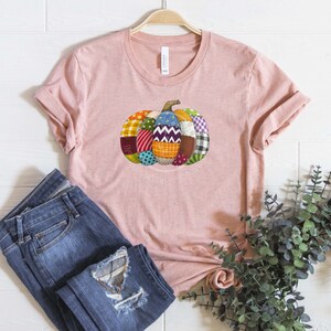 K&ouml;nnte beinhalten: Ein pinkfarbenes T-Shirt mit einem Patchwork-K&uuml;rbis-Design. Der K&uuml;rbis besteht aus verschiedenen Stoffen in den Farben Rot, Gr&uuml;n, Blau, Gelb und Lila.