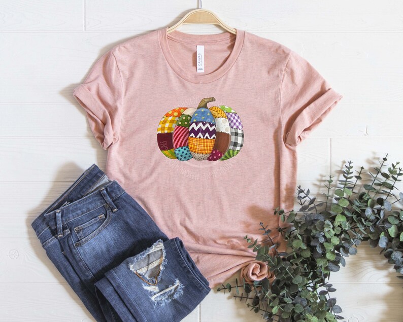 K&ouml;nnte beinhalten: Ein pinkfarbenes T-Shirt mit einem Patchwork-K&uuml;rbis-Design. Der K&uuml;rbis besteht aus verschiedenen Stoffen in den Farben Rot, Gr&uuml;n, Blau, Gelb und Lila.