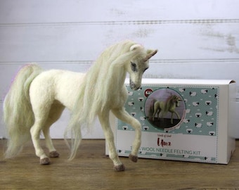 May include: A handcrafted, off-white unicorn figurine with a flowing mane and tail. The unicorn stands near a box labeled "Una" and "Wool Needle Felting Kit," featuring a picture of the unicorn. The kit includes materials for crafting the unicorn.