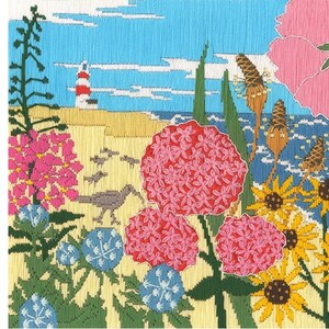 May include: Embroidered artwork showing a coastal scene with a lighthouse, beach, and ocean. The scene includes pink and yellow flowers, green foliage, and a bird. The sky is blue with white clouds.