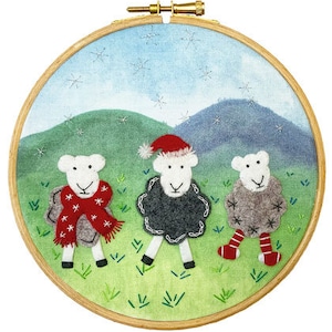 May include: A circular embroidery hoop with three sheep appliqués set against a green and blue landscape. One sheep has a red scarf, another a Santa hat, and the third wears red and white striped socks. The hoop is made of wood.