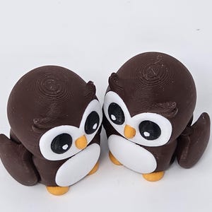 May include: Two brown owl figurines with white bellies, large black eyes, and yellow beaks and feet. The owls are 3D-printed and have a textured surface. They are positioned close together, suggesting a pair or a couple.