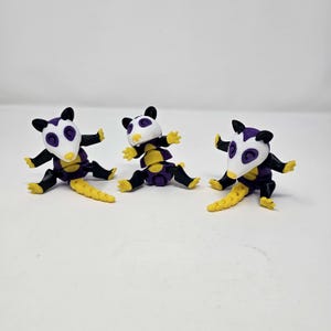May include: Three small, stylized opossum figurines. Each figurine has a white head with purple accents, black ears and limbs, a yellow and black body, and a yellow tail. The figurines are posed in a playful, seated position.