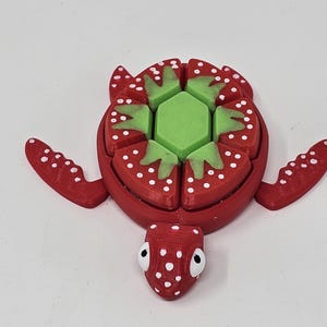 Op de afbeelding: Een rood en groen 3D-geprint schildpad speelgoed met een aardbeidesontwerp op zijn schild. Het schild is gemaakt van zes segmenten, elk met witte stippen en een groen bladerontwerp. Het hoofd van de schildpad is ook rood met witte stippen en heeft twee zwarte ogen.