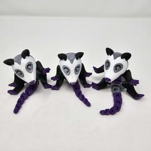 Può includere: Tre statuette di opossum stampate in 3D. Ogni statuetta ha una testa bianca con orecchie grigie, braccia e gambe nere e una coda segmentata viola. Gli opossum sono in posa carponi, con le code ricurve.