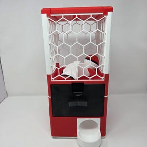 Op de afbeelding: Een rood-witte kauwgomballenautomaat met een zeshoekig kooi-ontwerp. De machine heeft een zwarte lade en een witte doseerbeker. In de kooi zitten witte en rode objecten.