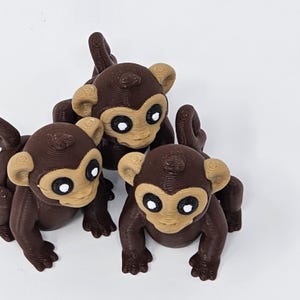 May include: Three brown monkey figurines with tan faces and ears, and large white eyes. The monkeys are in a playful pose, with their arms and legs extended. The figurines are likely 3D printed.