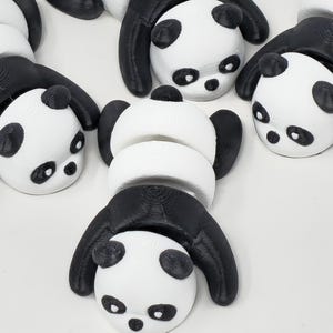 Peut inclure: Plusieurs jouets en forme de panda, blancs et noirs. Chaque panda présente une tête et un corps blancs, avec des oreilles, des yeux et des membres noirs. Les jouets sont disposés de manière ludique, mettant en valeur leur design unique.