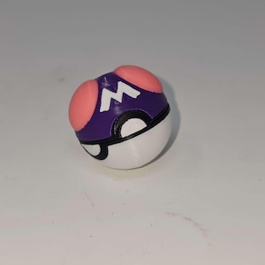 Puede incluir: Una Poké Ball impresa en 3D, de forma esférica. La parte superior es morada con una "M" blanca y dos círculos rosas. La mitad inferior es blanca, con una banda negra que separa las dos mitades. Un divertido objeto de colección.