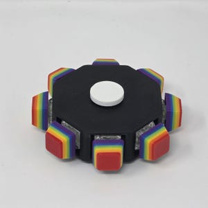 May include: A black fidget spinner with rainbow-colored edges and a white center button. The edges are square and have a gradient of colors, including red, orange, yellow, green, blue, and purple.