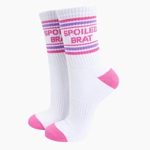 May include: A pair of white crew socks featuring pink toes and heels. The upper portion of the socks displays horizontal stripes in pink and purple, with the text "SPOILED BRAT" printed in pink capital letters between the striped sections.