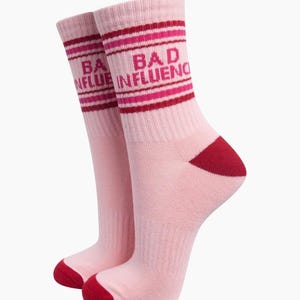 May include: A pair of light pink crew socks featuring dark red and bright pink horizontal stripes at the top. The text "BAD INFLUENCE" is printed in dark red lettering across the ribbed cuff area. The socks have a solid dark red heel and toe.