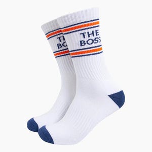 May include: A pair of white crew socks featuring dark blue toes and heels. The upper portion of the socks includes two horizontal orange stripes bordered by thin dark blue lines. The text THE BOSS is printed in dark blue on the outer side.
