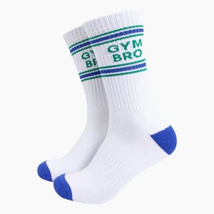 May include: A pair of white crew socks featuring blue and green horizontal stripes around the ribbed cuffs. The text GYM BRO is printed in green on the outer side of each sock. The toe and heel sections are accented with a solid blue colour.
