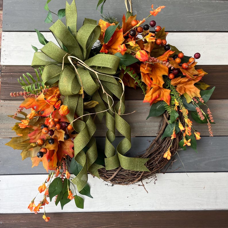 May include: A fall-themed wreath with a grapevine base, adorned with orange and yellow leaves, berries, and greenery. A large olive green ribbon adds a decorative touch. Perfect for autumn decor.