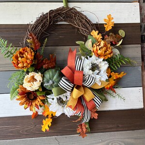 May include: A fall-themed wreath with a grapevine base, adorned with orange, white, and yellow sunflowers, chrysanthemums, and fall leaves. A green pumpkin and a multi-colored bow add to the festive design. The wreath is perfect for seasonal home decor.