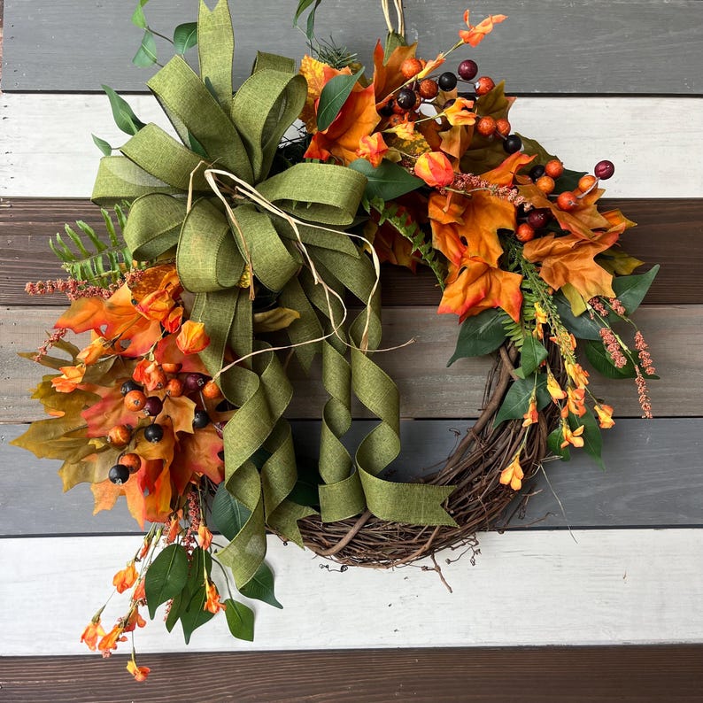 May include: A fall-themed wreath with a grapevine base, adorned with orange and yellow leaves, berries, and greenery. A large olive green bow with long, flowing ribbons adds a decorative touch. Perfect for autumn decor.