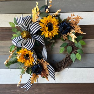 May include: A decorative wreath featuring sunflowers, navy blue flowers, and greenery. A navy and white striped ribbon is tied in a bow. The wreath is made of woven twigs and includes faux berries and leaves. The color palette includes yellow, navy, green, and brown.