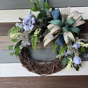 May include: A handcrafted Christmas wreath with a natural grapevine base. It features blue roses, green ferns, and frosted greenery. A large bow with "Merry Christmas" text and a Christmas tree pattern adds a festive touch. The wreath is perfect for holiday decor.