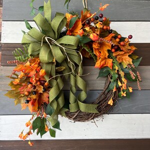 May include: A fall-themed wreath featuring a grapevine base adorned with orange and yellow autumn leaves, berries, and greenery. A large olive green ribbon bow adds a decorative touch to the top of the wreath. The wreath is displayed against a wooden background.